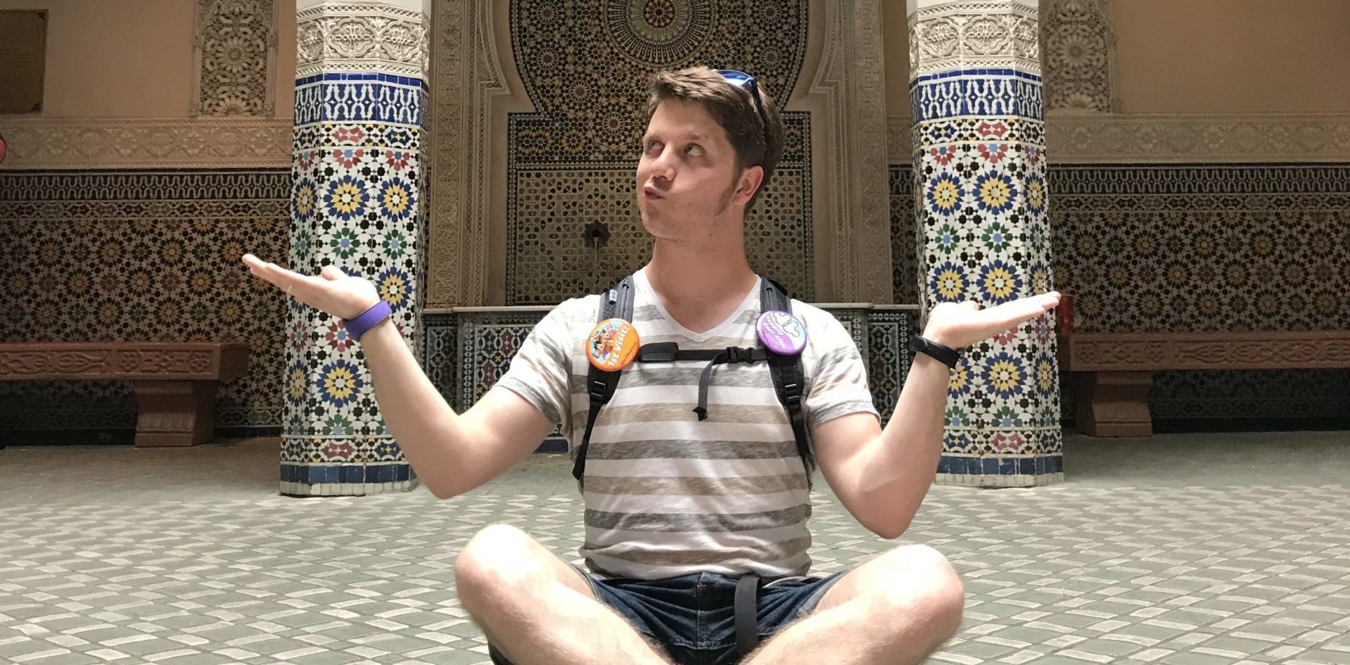 Ich im Schneidersitz vor einer orientalisch gestalteten Wand mit bunten Mosaiken. Die Hände halte ich neben meinen Schultern mit den Handflächen nach oben.