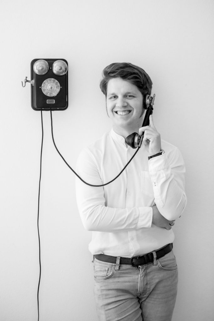 Martin an einer Wand neben einem alten Wandtelefon lehnend. Er hält den Hörer am Ohr und lächelt.